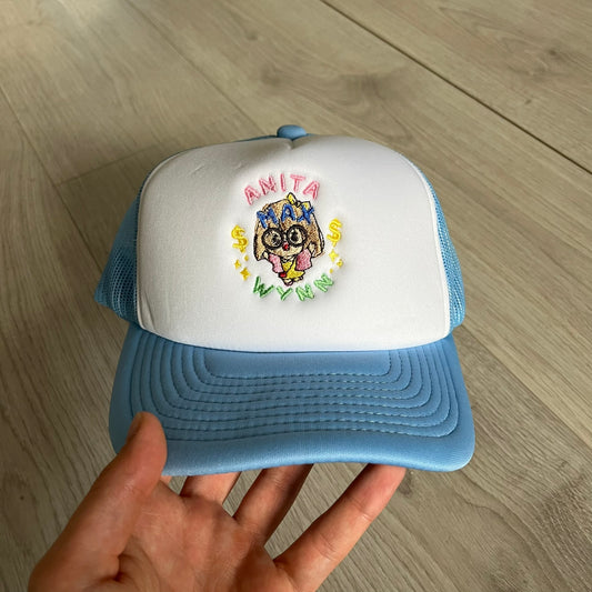 Drake Anita Max Wynn Pink Trucker Hat with a white front panel and baby blue sides and cap, featuring a collection of graphics including a woman with an oversized head, two yellow dollar signs, and “ANITA” in pink, “MAX” in blue, and “WYNN” in green. Designed with a mesh back for breathability, a snapback closure for adjustability, and polyester construction to keep the head cool. A playful tribute to Drake’s alter ego and love of g4mbling.