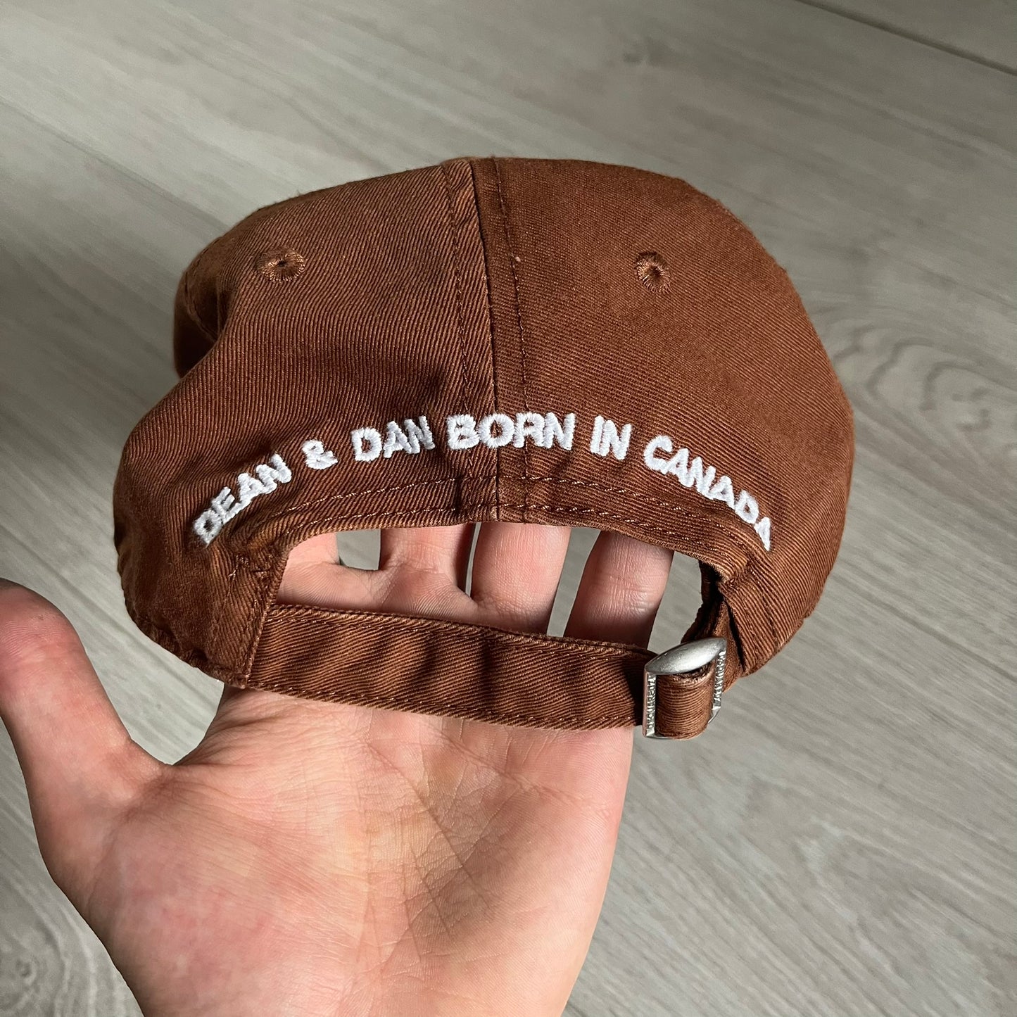 Brown cap with 'DEAN & DAN BORN IN CANADA' text held by a hand on a wooden surface