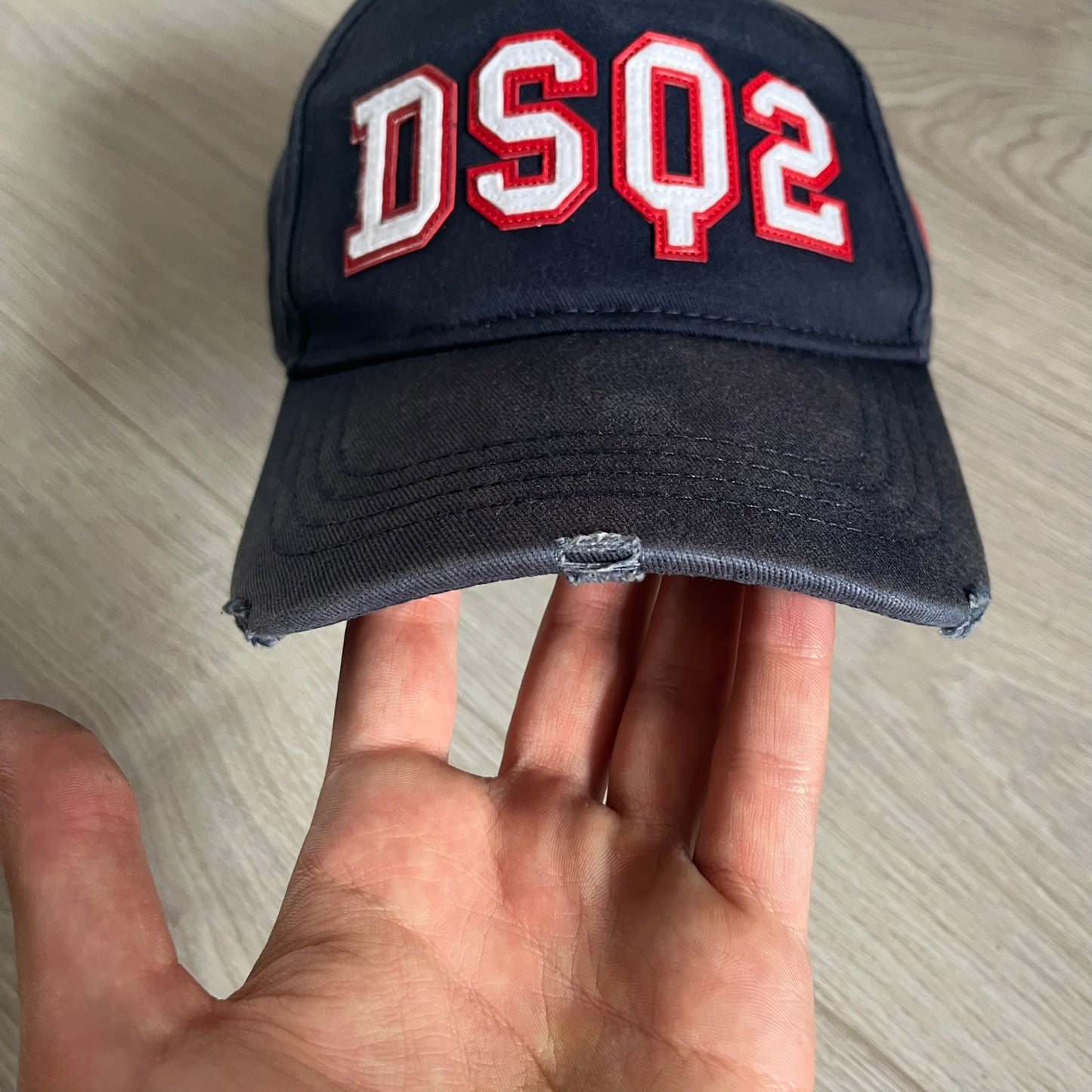 Black cap with 'DSQ2' in red and white held by a hand on a light wooden surface
