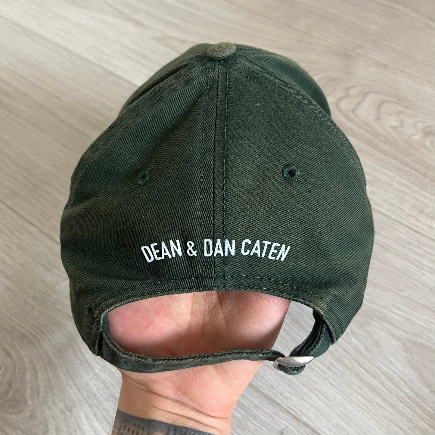 Green cap with 'DEAN & DAN CATEN' text held by a hand on a light wooden surface