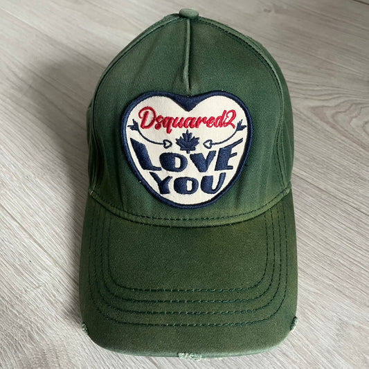 Green cap with a heart-shaped patch featuring 'Dsquared2 Love You' text on a wooden surface.
