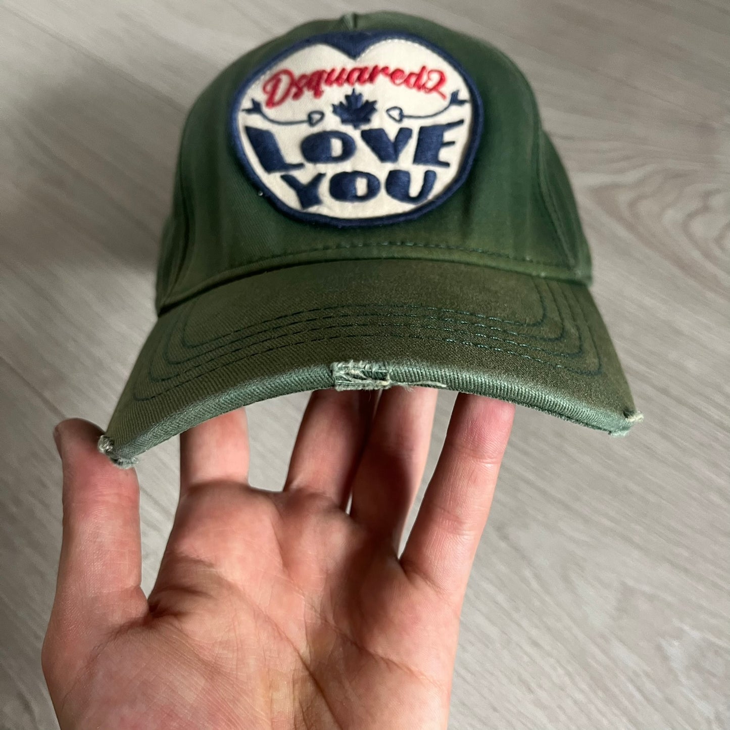 Green cap with 'Dsquared2 Love You' patch held by a hand on a light wooden surface