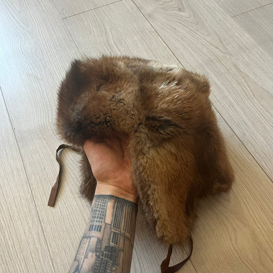 Fur ushanka hat featuring a Kulo tag inside and two leather tie strings. The front flap can be secured or folded back using small metal hooks for adjustable coverage. Designed for warmth and comfort, this classic winter piece offers both functionality and timeless style.