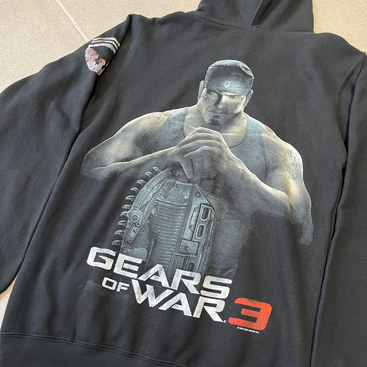 Gears of War 3 Marcus Fenix 2010 Promo Hoodie is a black zip-up hoodie with a dark Crimson Omen logo in orange tones on the left upper chest and a bloody Delta-One logo on one sleeve. The back features a large Marcus Fenix graphic with “Gears of War 3” text below it and a small 2010 Epic Games licensing mark.
