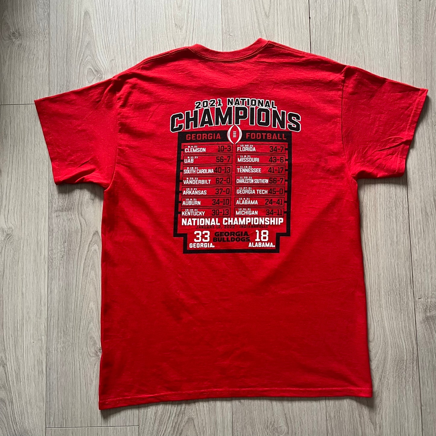 Red t-shirt with '2021 National Champions' text and game scores on a wooden surface