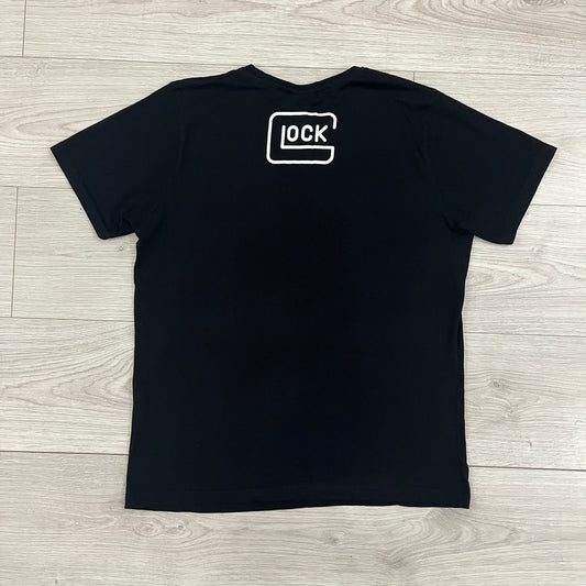 Glock Safe Action Pistol T-Shirt in black, made on an Orijin blank from 100% cotton. Features a large Glock pistol graphic on the front set inside a yellow circle, with “Glock Safe Action” text and Glock logo running around the graphic. The back has a white Glock logo printed below the collar. 