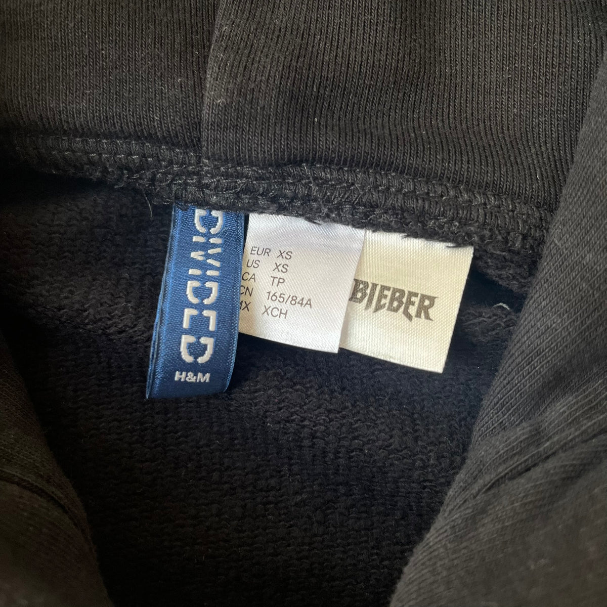 H&M 2016 Justin Bieber Purpose World Tour Black Hoodie is from the 2016 H&M Divided collaboration collection, featuring a white "Purpose" logo with "World Tour" text below it on the front. Both sleeves display big red "WORLD TOUR" text.