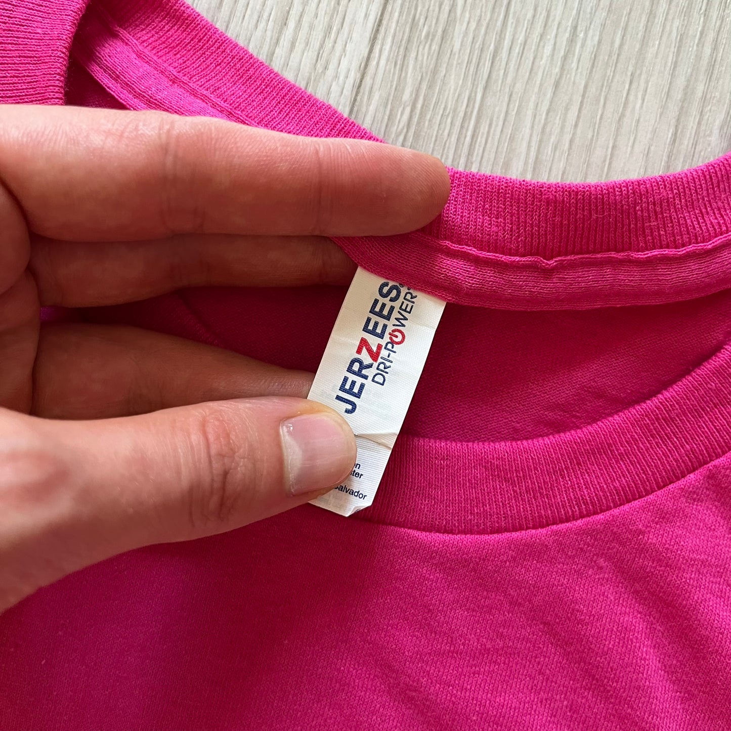 Hand holding a pink garment with a Jerzees tag on a light wooden surface
