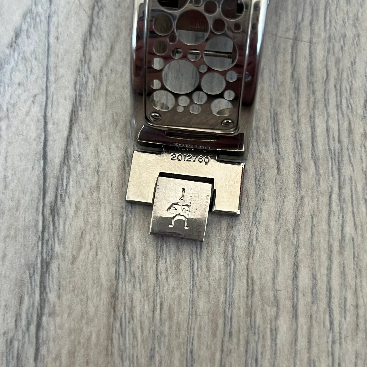 Hi-Tek Alexander 90s Perforated Analog Watch is a Hi-Tek London watch featuring a black dial with orange hands. The hour markers are represented by Hi-Tek figure graphics instead of traditional numerals. The dial is covered by a metal protective plate with a perforated hole pattern. Around the case is a metal ring with Hi-Tek London branding and a figure detail. The bracelet consists of two rigid metal sections with alternating small and large perforations.