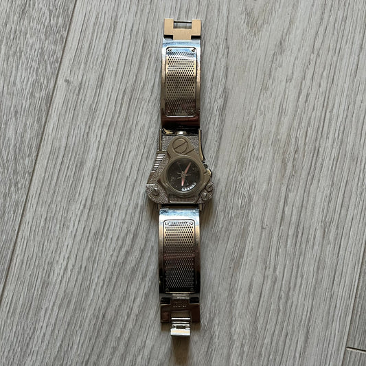 The Hi-Tek Alexander “The Engine” analog watch is a 1990s model made in England, designed with a sculptural case shaped like a motorcycle engine. It features a black dial with Alexander Hi-Tek figure logos in place of hour markers, pink hour and minute hands, and a Hi-Tek figure logo used as the seconds hand. A small sub-section at the bottom displays the day in numeric form, while visible screws and textured detailing are integrated into the engine-style face. The watch has an alloy case with a stainl