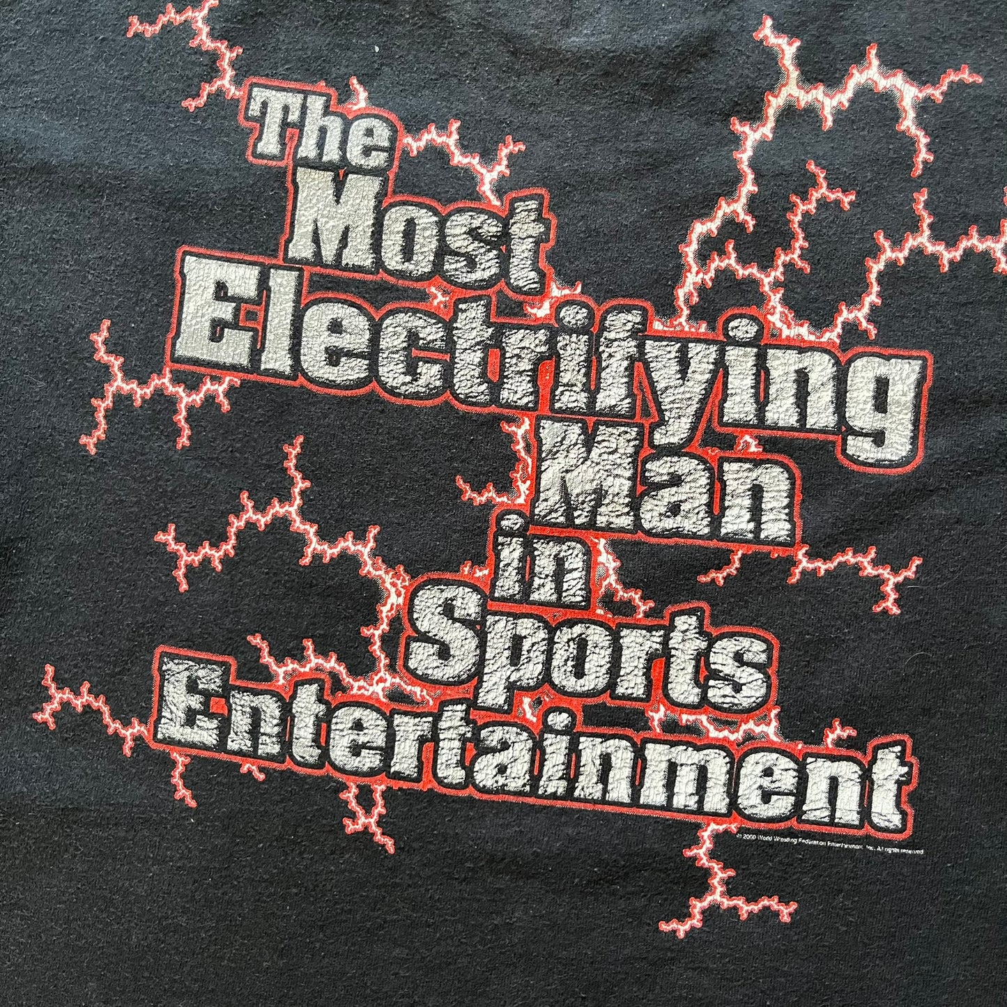 The Rock “Most Electrifying Man” 2000 Tee is a vintage WWE shirt featuring bold metallic-textured lettering with red lightning accents. The front proudly displays “The Rock,” while the back proclaims “The Most Electrifying Man in Sports Entertainment.” Licensed by World Wrestling Federation (WWF), this 2000 release is a must-have collectible for wrestling fans!