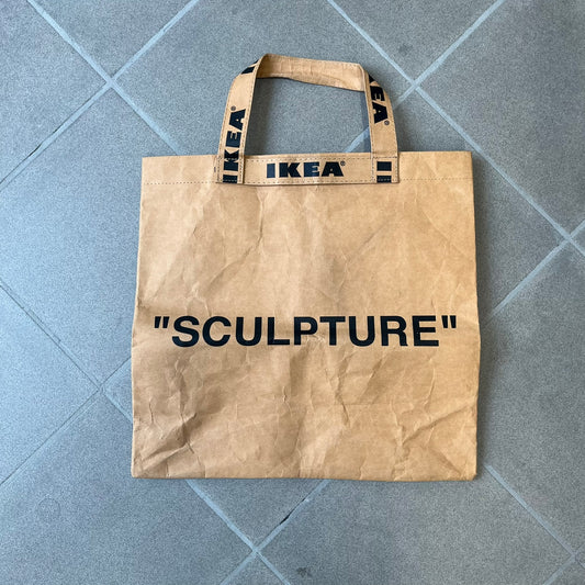 Ikea x Virgil Abloh Markerad "Sculpture" Carrier Bag from FW19 reimagines the iconic blue Frakta tote in reinforced brown paper. Embellished with Abloh’s signature quotation marks, the “SCULPTURE” print transforms a utilitarian object into conceptual design. A collectible symbol of functional art and everyday irony.