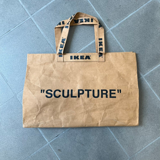 Ikea x Virgil Abloh Markerad "Sculpture" Carrier Bag from FW19 reimagines the iconic blue Frakta tote in reinforced brown paper. Embellished with Abloh’s signature quotation marks, the “SCULPTURE” print transforms a utilitarian object into conceptual design. A collectible symbol of functional art and everyday irony.