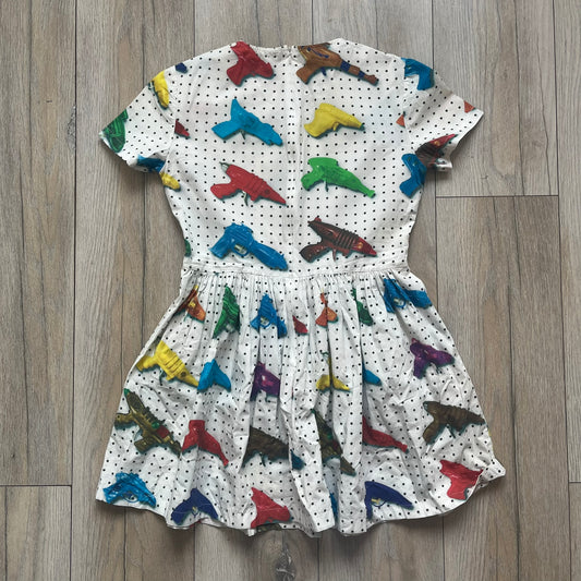 Jeremy Scott SS2016 Watergun Dress is a white flared short dress featuring an all-over print of multicolored realistic waterguns in varying sizes and orientations. The design is set against a black dotted pattern layered across the fabric. It has a fitted upper section with a flared skirt silhouette and includes a black Jeremy Scott neck tag.