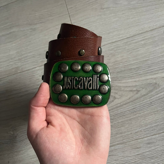 Just Cavalli Green Metal Buckle Studded Belt featuring a shiny green metallic buckle with the Just Cavalli logo in the center, surrounded by metallic studs. The brown leather strap is accented with a line of metallic studs along its length.