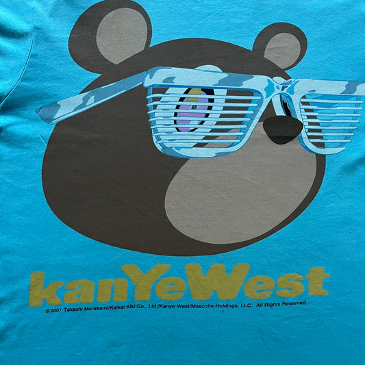 Kanye West 2012 Takashi Murakami 'Glow In The Dark Tour' Tee in light blue features the iconic bear with shutter glasses on the front. Below the graphic is yellow "kanYeWest" text and the 2007 Takashi Murakami/Kanye West licensing. Official merchandise from the 2012, this collectible tee includes the neck tag with Kanye logo and Mascotte Holdings licensing.