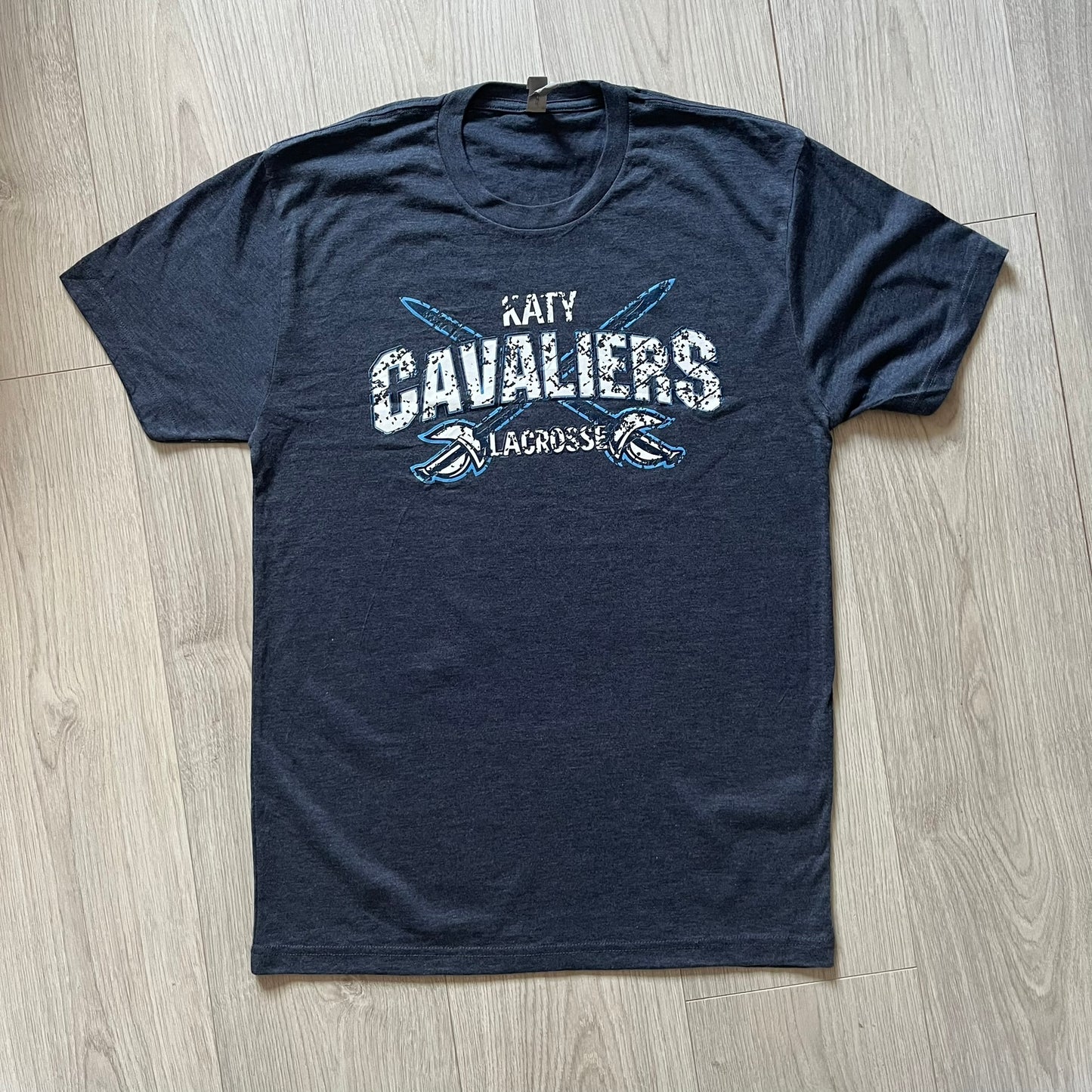 Navy blue t-shirt with 'Katy Cavaliers Lacrosse' text on a wooden floor