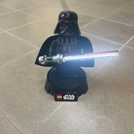 LEGO 2012 Darth Vader LED Desk Lamp (LGLLP2B) features a poseable oversized Darth Vader figure standing on a specialized base and holding a 12-LED lightsaber that functions as the light source. The helmet can be removed and the figure can be detached from the base, while the base has a LEGO Star Wars logo on the front and licensing details underneath; additional LEGO Star Wars licensing appears on the back of Vader. Powered by 3 AA batteries with a 30-minute auto-off timer. The original cape has been replac