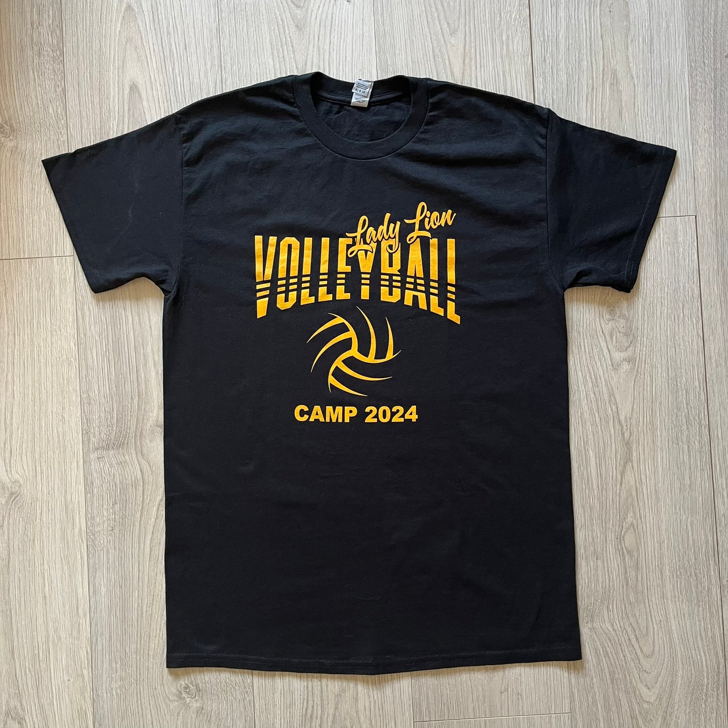 Black t-shirt with yellow 'Volleyball Camp 2024' text and volleyball graphic on a wooden surface