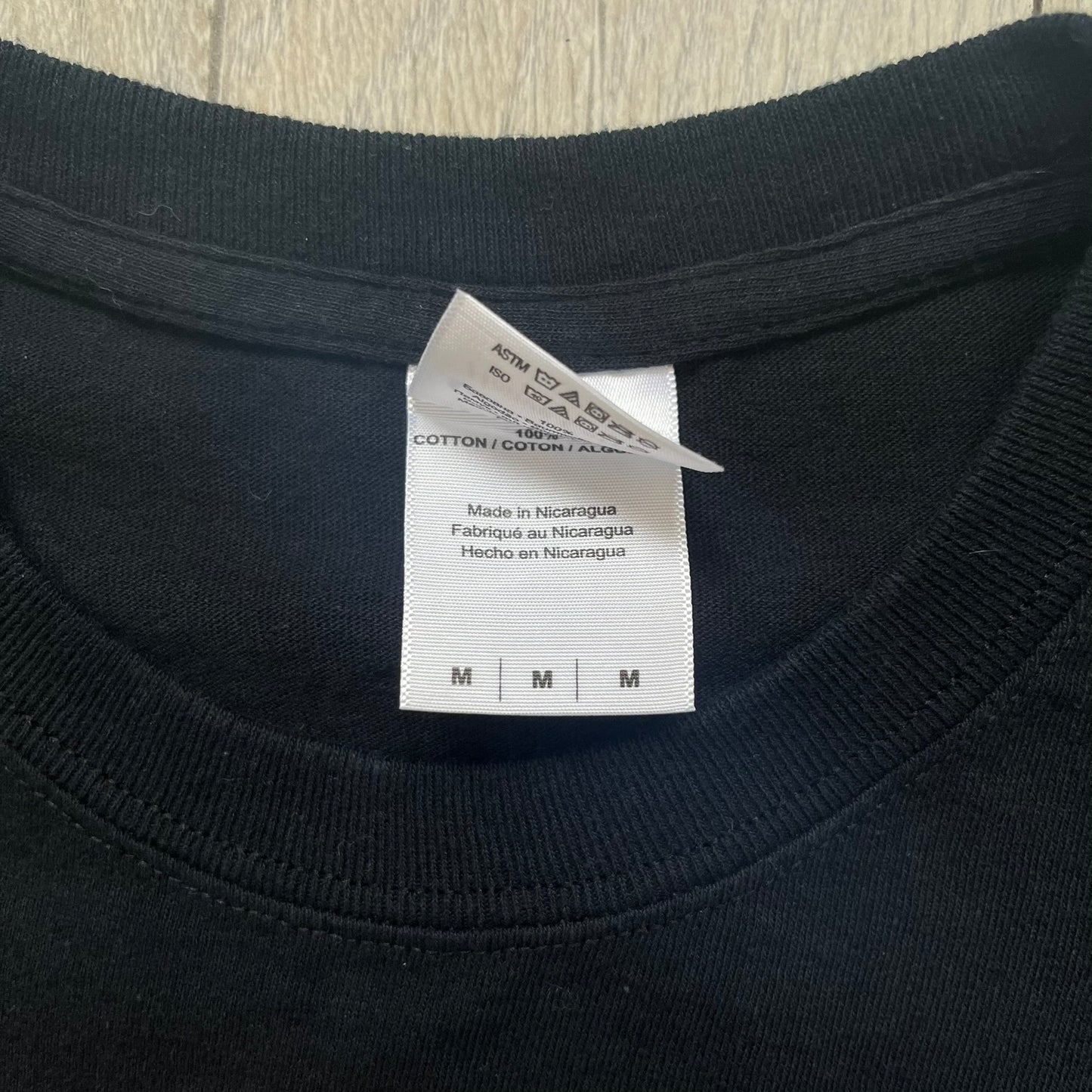 LEGO Minifigures “New Series” 2015 Tee is a black T-shirt featuring front and back prints. The front displays “LEGO Minifigures” text above eight characters arranged in a circle, with 2015 LEGO licensing and “New Series” text below. The back repeats the logo with a yellow minifigure in the center and “ASK ME” text underneath, likely produced as an employee promotional item.