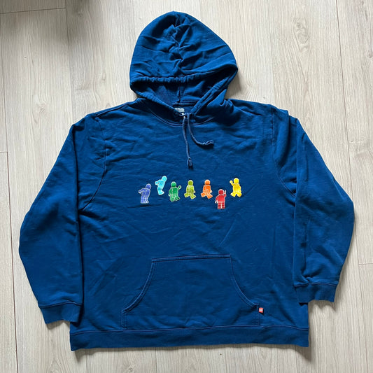 Lego Rainbow Minifigures Blue Hoodie from 2010, officially licensed by the Lego Group. Crafted from 100% cotton, it features seven colorful Lego minifigures in motion across the chest and a single red figure waving on the upper back. Designed in a bright blue colorway with a small Lego tag by the front pocket.