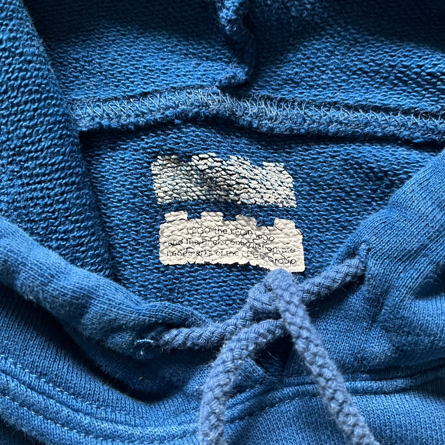Lego Rainbow Minifigures Blue Hoodie from 2010, officially licensed by the Lego Group. Crafted from 100% cotton, it features seven colorful Lego minifigures in motion across the chest and a single red figure waving on the upper back. Designed in a bright blue colorway with a small Lego tag by the front pocket.