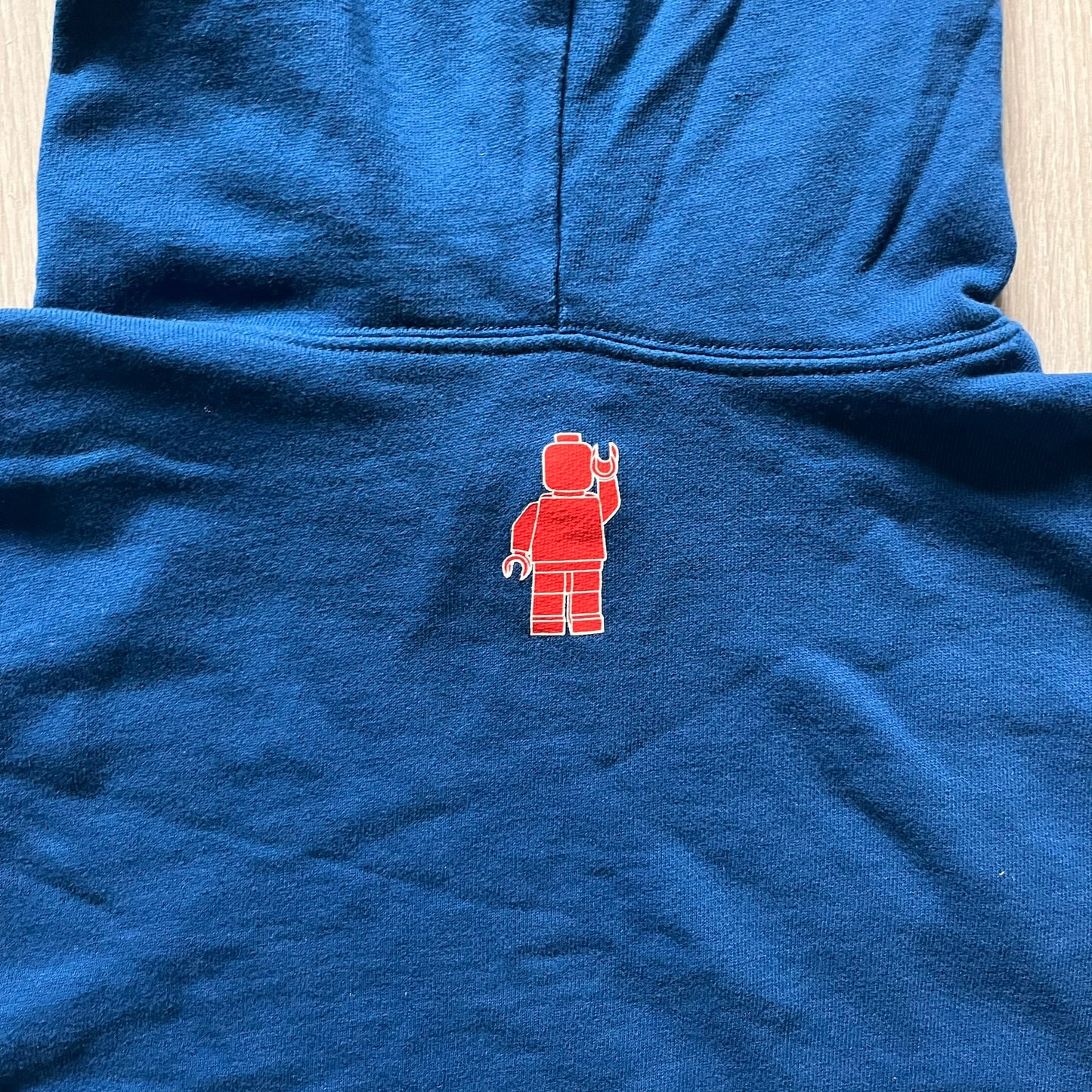 Lego Rainbow Minifigures Blue Hoodie from 2010, officially licensed by the Lego Group. Crafted from 100% cotton, it features seven colorful Lego minifigures in motion across the chest and a single red figure waving on the upper back. Designed in a bright blue colorway with a small Lego tag by the front pocket.
