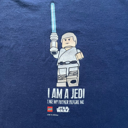 Vintage 2007 Lego Star Wars tee featuring Luke Skywalker with a lightsaber and the quote “I am a Jedi like my father before me.” Small Lego Star Wars logo sits underneath the graphic. Printed on a classic navy Fruit of the Loom blank perfect for collectors and SW fans.
