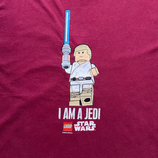 This Lego Star Wars 2008 Jedi Tee (Medium) in burgundy showcases Luke Skywalker wielding a blue lightsaber with a 'cracked' “I Am a Jedi” text. Officially licensed by Lego Star Wars, the tee includes authentic 2008 branding under the Luke minifigure graphic.