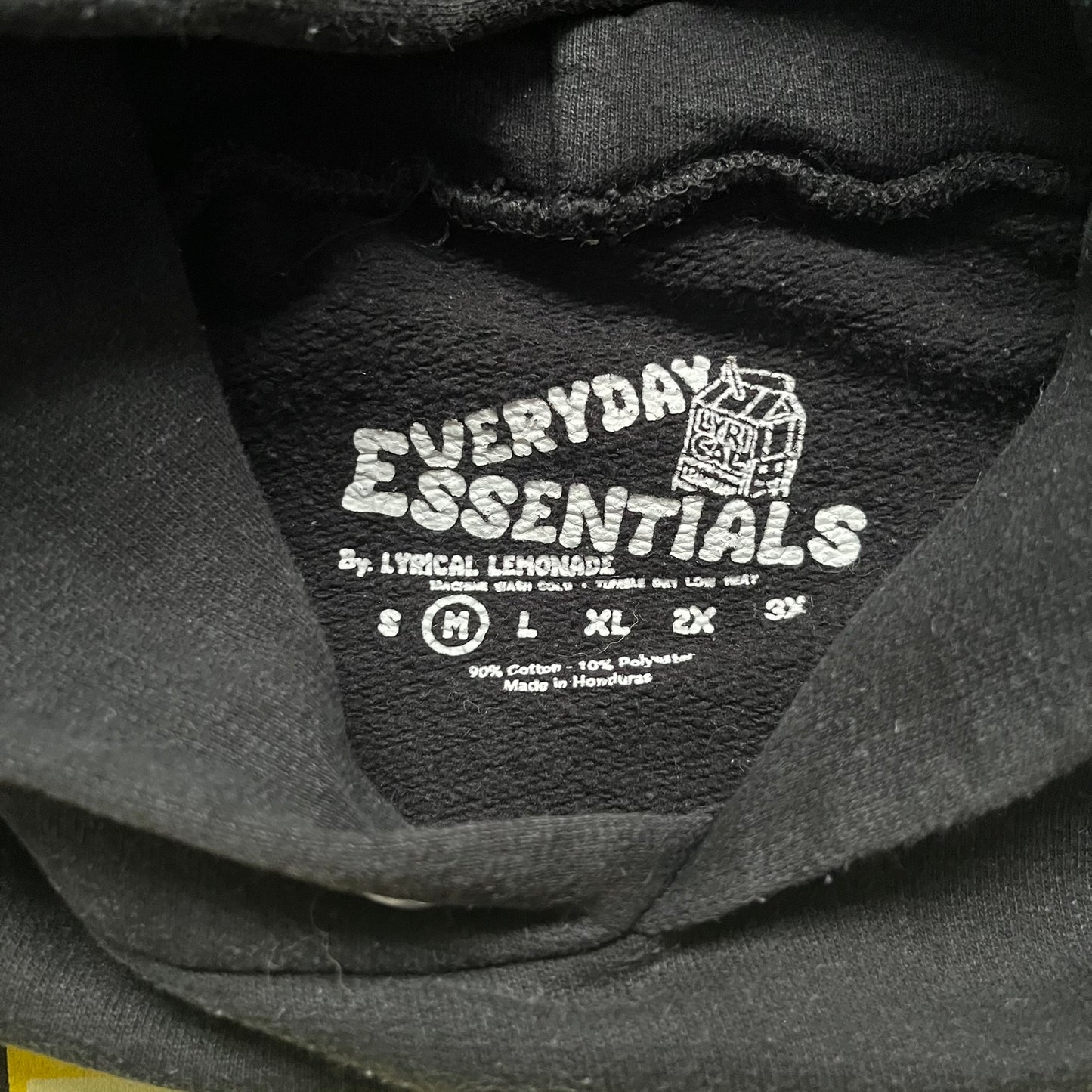 Lyrical Lemonade Everyday Essentials Yellow Logo Hoodie is a black pullover hoodie featuring a large two-tone yellow Lyrical Lemonade logo printed on the front. It includes an Everyday Essentials neck tag and classic comfortable hoodie construction.