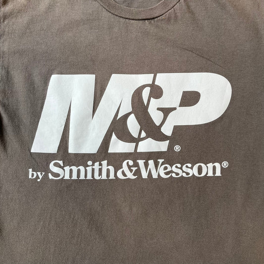 M&P by Smith & Wesson Gray Tee features a washed grey finish with a large M&P logo printed across the front and smaller "by Smith & Wesson" text underneath. Made from 100% cotton on a Gildan blank