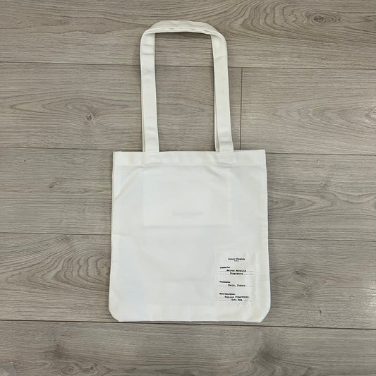 Maison Margiela Replica Tote Bag in off-white with a textured finish inspired by natural jute. Features “Replica” fragrances patch on one side, an interior slip pocket, and small “Maison Margiela Paris” text on the other side. Made in Paris, versatile for daily errands, work, or casual outings.