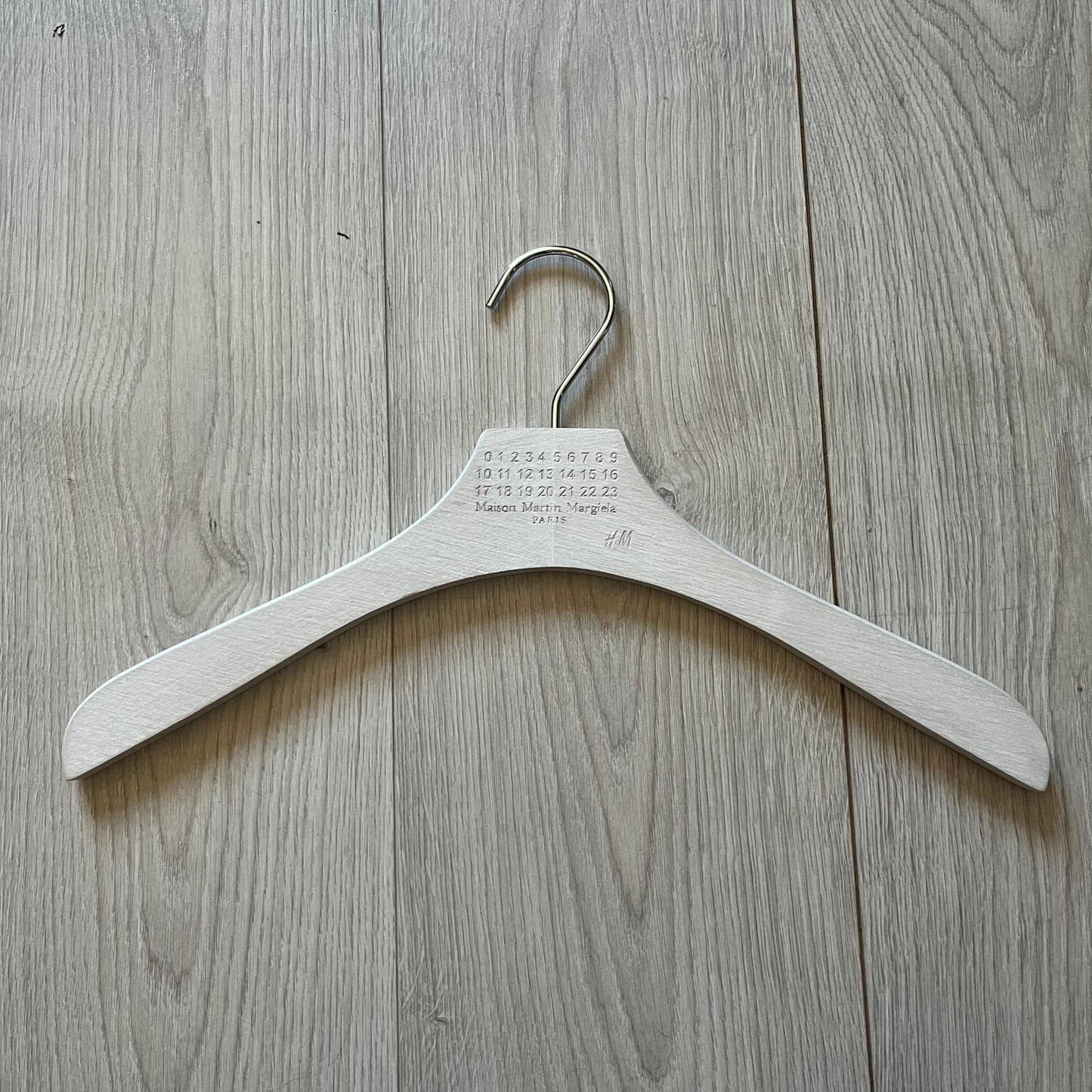 Maison Margiela x H&M 2012 clothing hanger in a white-washed wood finish. Features a carved Margiela and H&M logo with a clean, classic silhouette. Ideal for displaying tees, hoodies, or jackets with a touch of designer minimalism.