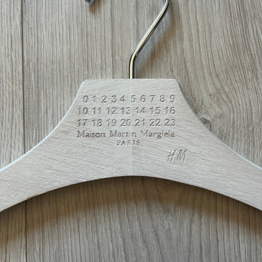 Maison Margiela x H&M 2012 clothing hanger in a white-washed wood finish. Features a carved Margiela and H&M logo with a clean, classic silhouette. Ideal for displaying tees, hoodies, or jackets with a touch of designer minimalism.