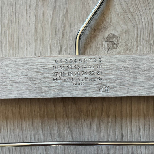 Maison Margiela x H&M 2012 hanger in a white-washed wood finish with engraved logos. Features a straight wooden bar with metal clips, perfect for hanging pants, skirts, or shorts. A rare, functional collectible from this iconic collaboration.