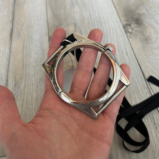 Maison Margiela x H&M AW09 Enlarged Watch Frame Bracelet is a stainless steel bracelet from the 2012 collaboration between Maison Margiela and H&M, based on an original Autumn/Winter 2009 design. The piece is shaped like an oversized watch case but functions as a bracelet, featuring a side clasp opening mechanism. It includes engraved tonal branding on the interior and can also be worn as a pendant. Made in Italy, it comes with the original tag.
