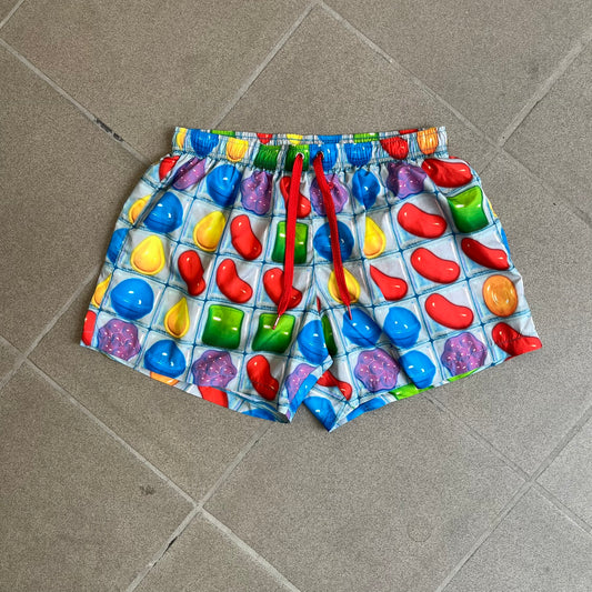 Moschino Candy Crush 2017 Swimming Shorts feature an all-over print inspired by the Candy Crush game board, with colorful candy graphics covering the fabric. The back is detailed with a large Moschino logo in a gold Candy Crush-style font. The shorts include red drawstrings with Moschino-branded metal aglets, a mesh inner lining, and are made in Italy.