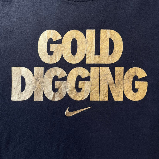 Nike 2012 Olympics “Gold Digging” Black Tee features a black slim-fit T-shirt with gold “Gold Digging” text on the front and a small Nike logo underneath. The design was released in connection with the 2012 Olympics and was later discontinued following controversy surrounding the wording.
