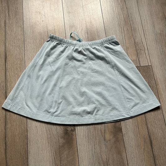 Nike 90’s Challenge Court Tennis Skirt is a baby blue women’s tennis skirt made in Thailand. It features a small green and red Nike Court logo patch at the lower hem, a Dri-FIT patch at the top, and an adjustable waistband with drawstrings. 