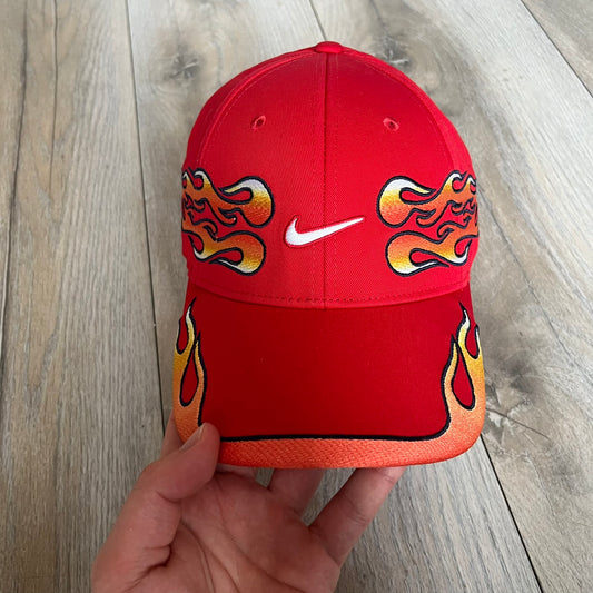 Nike Club Semi-Structured OG University Red Flame Cap is a red baseball cap featuring a white Nike Swoosh centered on the front panel. Embroidered flame graphics in orange, yellow, and red extend across the front of the visor and side panels. A small white Nike Swoosh is placed at the back. The design is identified by code IH9258-657. 