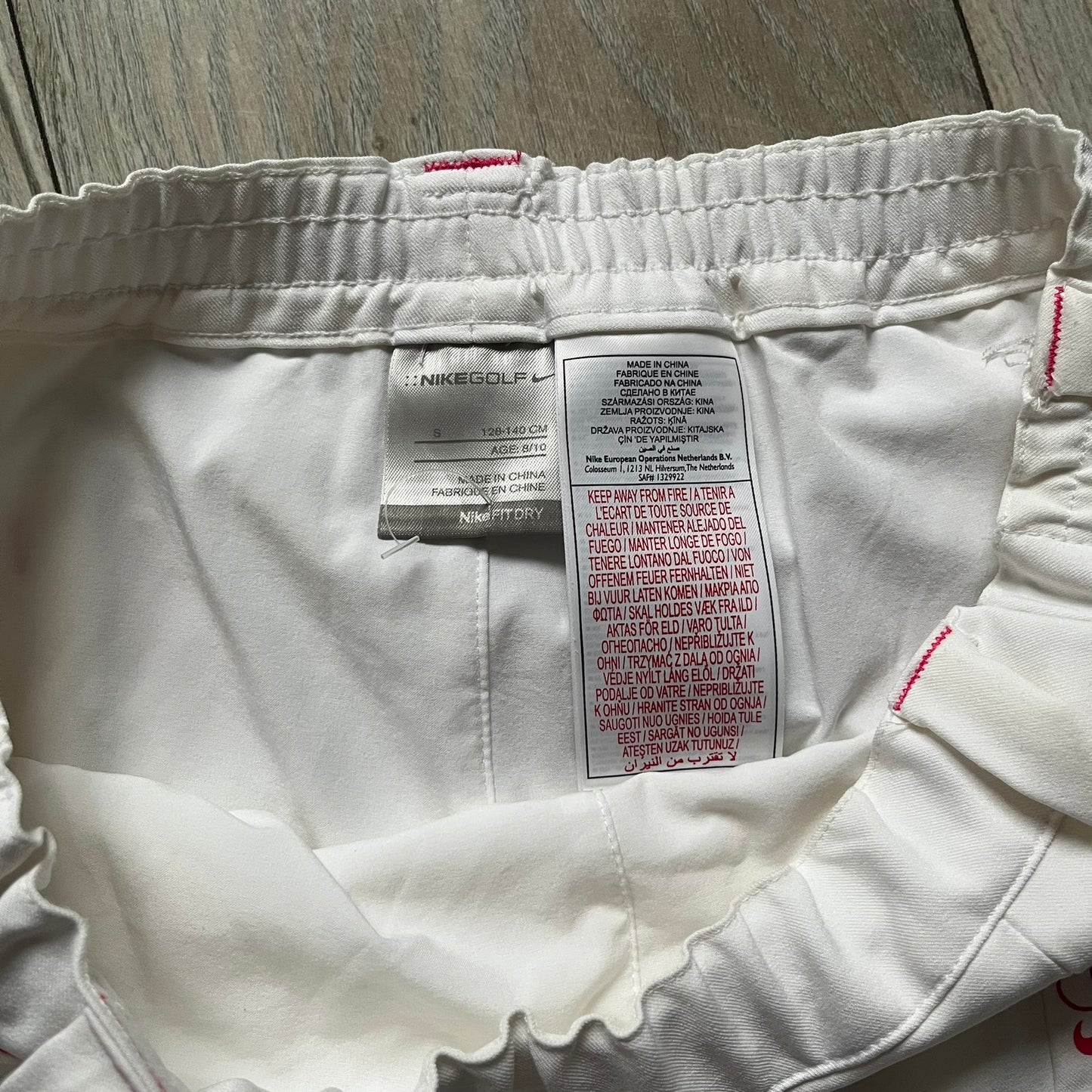 Nike Fit Dry White/Pink Golf Skirt is a women’s golf skort with built-in inner shorts. The white skirt features belt loops at the waistband, front pockets, a small back pocket, and side cutouts on both sides for flexibility. Below the left front pocket there is a small pink floral-style detail along with a Nike Golf patch.