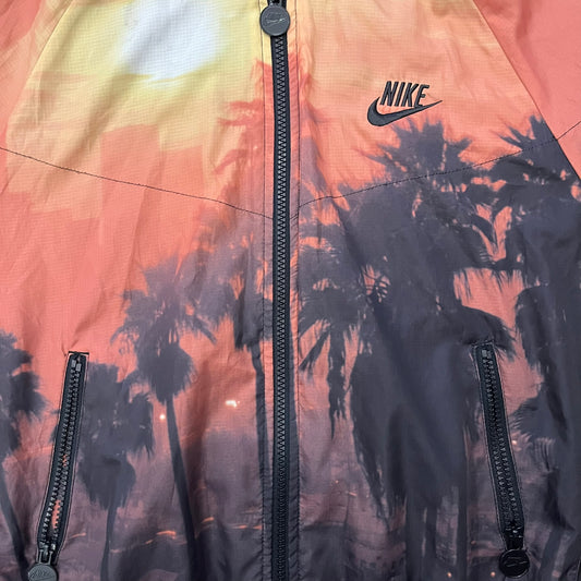 Nike “Beach Sunset” Split Windbreaker WMNS features dual zippers along the front and back, allowing the jacket to separate and merge with other compatible models from the collection. The all-over print shows an orange and yellow sunset with black palms, complemented by black hems and interior lining. Finished with circular metal Nike zip pulls and a small black embroidered Swoosh on the right chest.