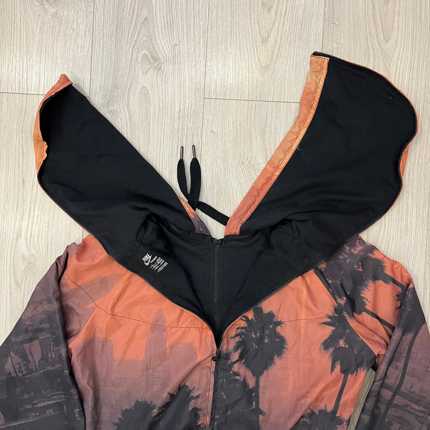 Nike “Beach Sunset” Split Windbreaker WMNS features dual zippers along the front and back, allowing the jacket to separate and merge with other compatible models from the collection. The all-over print shows an orange and yellow sunset with black palms, complemented by black hems and interior lining. Finished with circular metal Nike zip pulls and a small black embroidered Swoosh on the right chest.