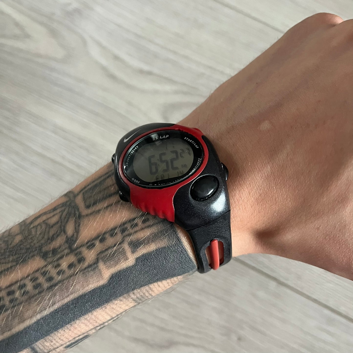 Digital watch with red and black strap on a wrist with tattoos, on a light wooden surface.