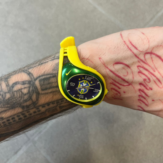 Nike Triax Blaze Brazil WK0008 Analog Watch is a vintage watch featuring a yellow rubber strap, green metal case, and blue metallic dial. The dial displays a large full-color Brazil Football crest with white main numerals, while a yellow Nike Swoosh is positioned at the top of the case. The watch is water resistant up to 50 meters and is constructed from stainless steel and plastic materials.