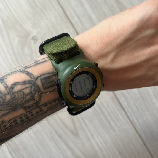 Green digital watch with a Nike logo on a wrist with tattoos, on a light wooden surface.