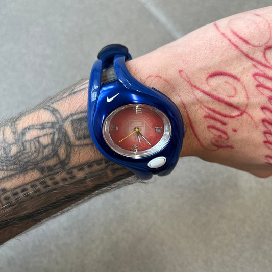 Nike Triax Swift 'Manchester United' WR0078 Blue Analog Watch featuring blue straps and a matching blue outer shell covering the metal case. The inner case is metallic blue with a white light button and a Nike Swoosh on the top. The dial has a light red gradient finish with a large Manchester United crest in the center. Analog display with an integrated backlight that illuminates the dial.