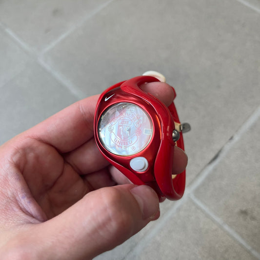 Nike Triax Swift 'Manchester United' WR0078 Red Analog Watch featuring red straps and a matching red outer shell covering the metal case. The inner case is metallic red with a white light button and a Nike Swoosh on the top. The dial has a light red gradient finish with a large Manchester United crest in the center. Analog display with an integrated backlight that illuminates the dial.