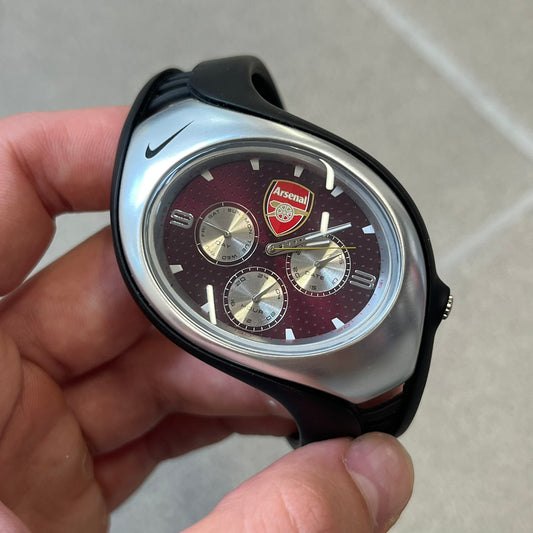 Nike Triax 'Arsenal' Swift 3i WR0091 Analog Watch with black rubber straps and a silver-tone case featuring a black Nike Swoosh at the top. The dark red metallic dial has a recessed dotted texture, Arsenal F.C. crest at the top and three silver subdials for day, date, and hour. Seconds markings are printed on the inner edge of the case and are visible only at an angle. 