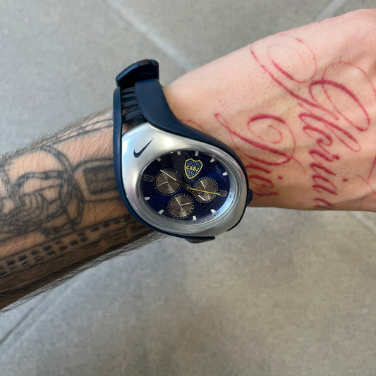 Nike Triax 'Boca Juniors' Swift 3i WR0091 Analog Watch with black rubber straps and a silver-tone case featuring a black Nike Swoosh at the top. The navy metallic dial has a recessed dotted texture, Boca Juniors crest at the top and three silver subdials for day, date, and hour. Seconds markings are printed on the inner edge of the case and are visible only at an angle. 